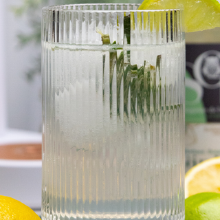 Verre de mocktial Bota Fiiz sans Alcool en premier plan avec des demi citrons et demi limes. En arrière-plan, une machine à eau pétillante Indigo et une bouteille de gin bota Sobr. 